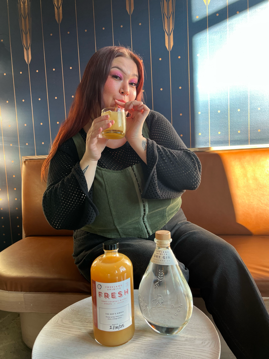 Grace Aasen of the Oregon non-profit the Cupcake Girls sipping on a Bee's Knees cocktail . On a table is a bottle of Bee's Knees seasonal FRESH cocktail mixer and a bottle of Freeland Dry Gin.